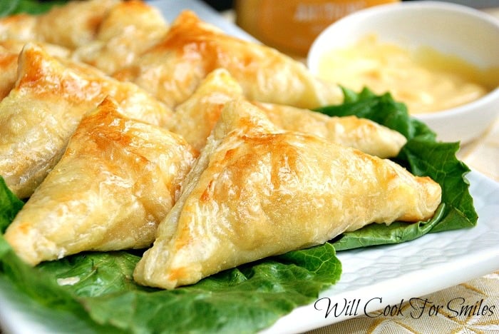 8 triangular shaped honey dijon chicken pockets on a bed of lettuce on white plate with a small sauce bowl in background filled with honey dijun dressing