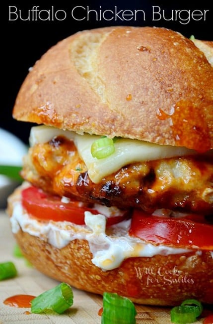 Buffalo chicken burger with cheese tomatoes and sauce on a bun on a cutting board with green onions 