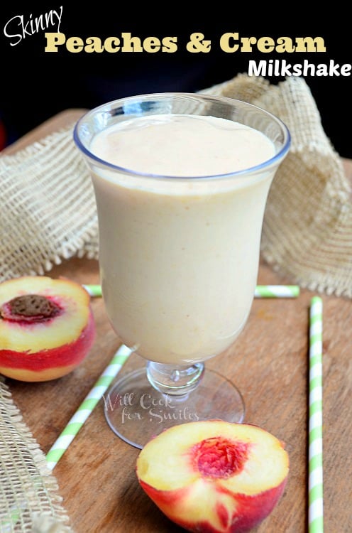 dessert glass filled with skinny peaches and cream milkshake on wooden table with green and white straws and peaches surrounded by burlap cloth