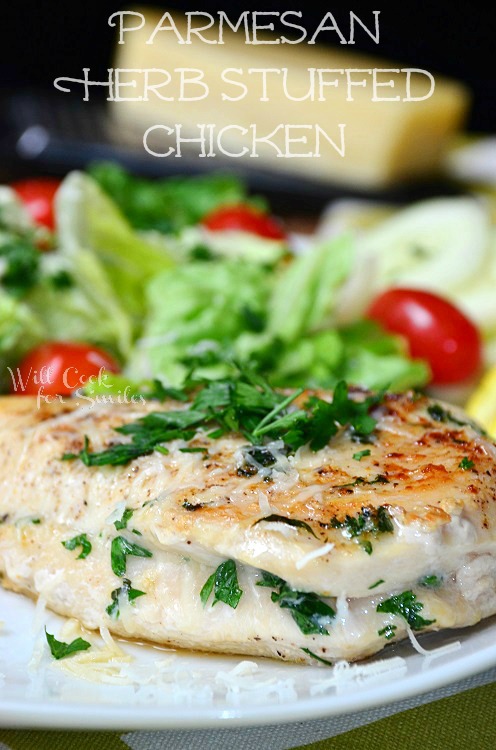 Parmesan and Herb Stuffed Chicken is plated on a white dish. Extra herbs are on top of the chicken. A salad with red tomatoes accompanies the dish.