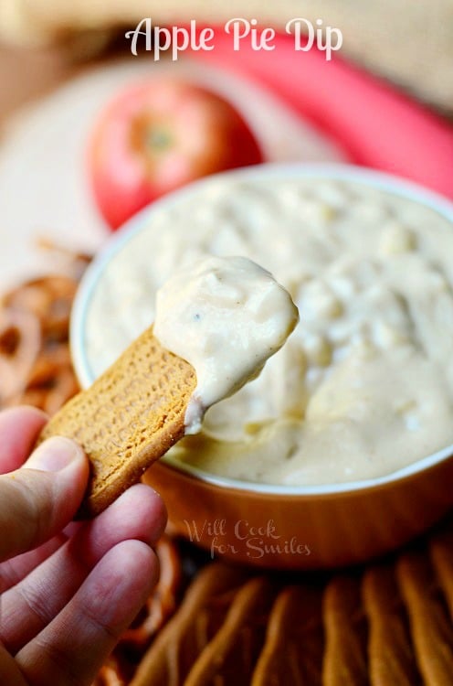 hand holding cookie with dip on it above brown bowl filled with apple pie dip surrounded by cookies and pretzels