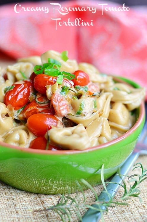 green and red clay bowl filled with creamy rosemary tomato tortellini on a burlap mat with fork to the bottom right