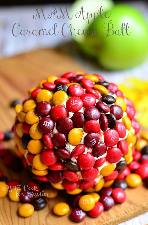 dessert cheese ball covered in M&Ms on wood cutting board with apple in background to the left