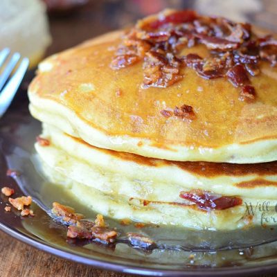 stack of maple bacon pancakes on black plate with fork to the left and syrup jar in background to the left