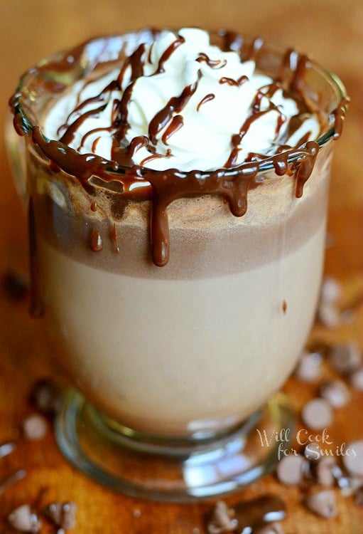 Hot Chocolate in a glass with whipped cream and chocolate sauce on a wood table with chocolate chips around it 