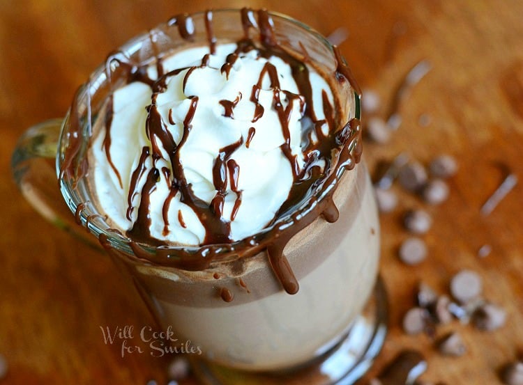 view from above of clear glass coffee mug filled with adult hot chocolate with whipped topping on a brown table with chocolate chips scattered around the bottom of the glass