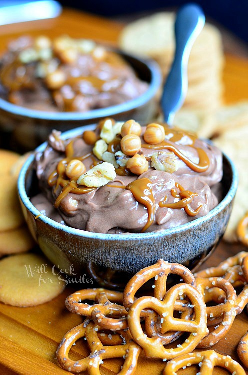 2 blue and black clay bowls filled with chocolate turtle dip with pretzles and cookies scattered around bowls and hershey's chocolate in glass bottles