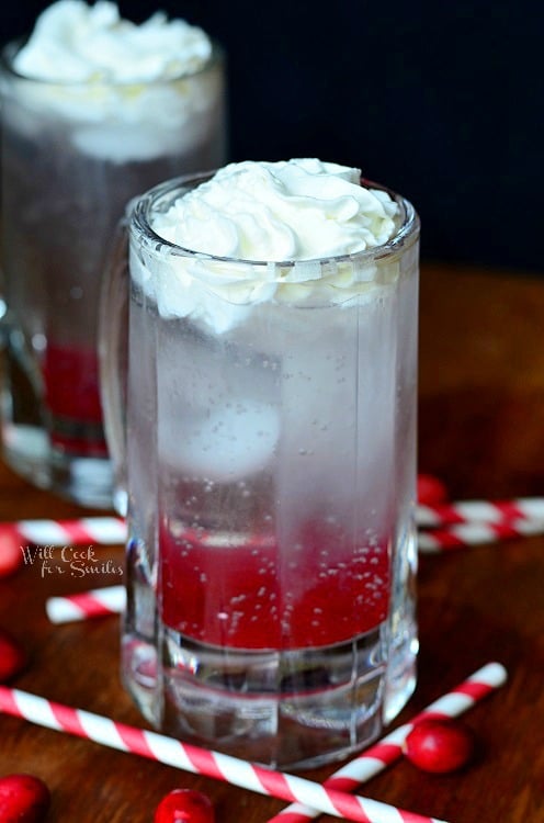 Cranberry Italian Soda in a glass mug with whipped cream in top and red and white straws around the bottom on the wood table