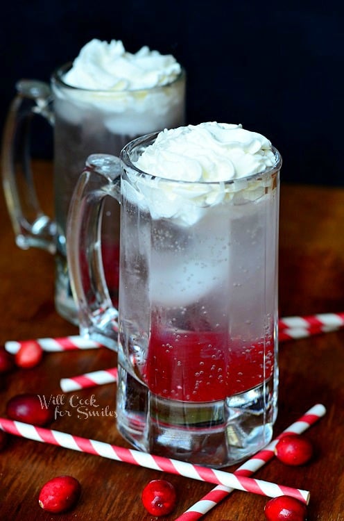 2 beer mugs filled with cranberry italian soda on wood cutting board with straws and cranberries scattered around mugs