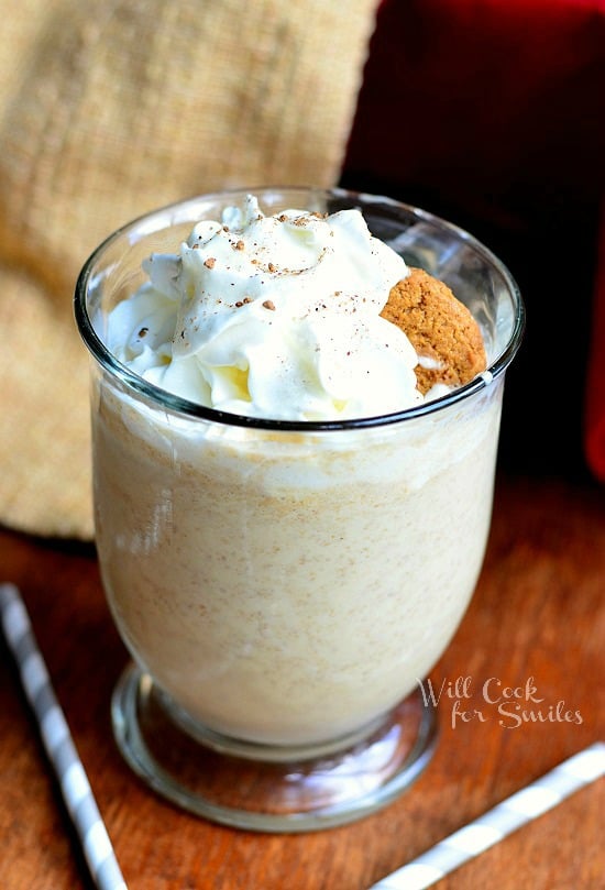 1 glass dessert mug with handle holding gingersnap milkshake with whipped topping on a wood table with strawas scattered around glass and brown cloth in background to the left