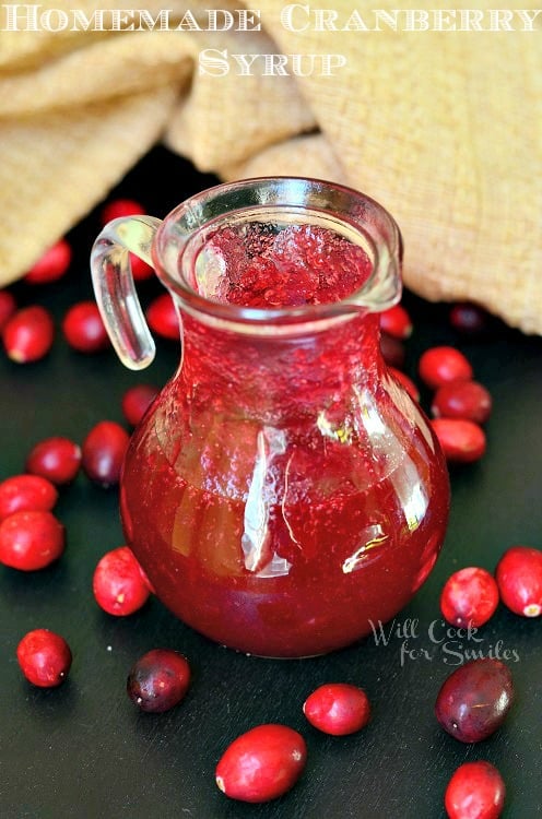 glass syrup jar filled with cranberry syrup on a black table with cranberries scattered around jar and brown tablenapkin in background at the top of the picture