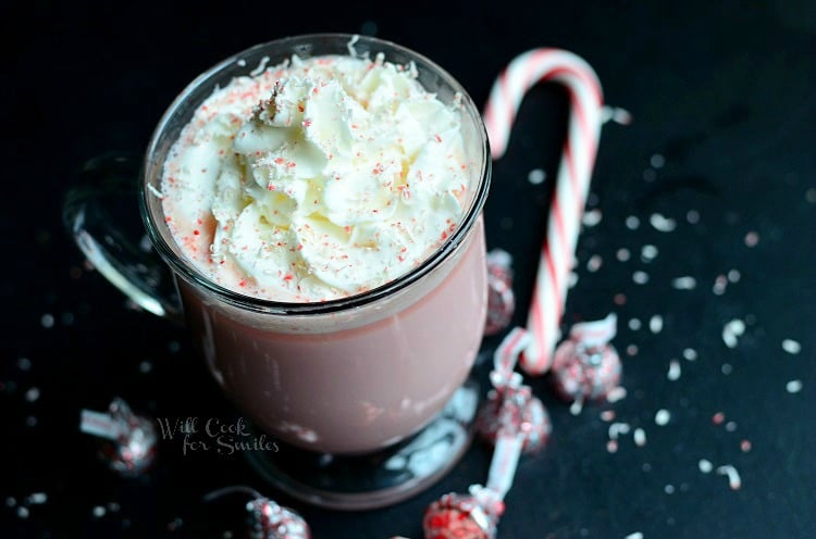 above view of clear glass coffee mug filled with peppermint white chocolate hot cocoa topped with whipped topping as peppermint hershey kisses and a candy cane are scattered at bottom of coffee mug