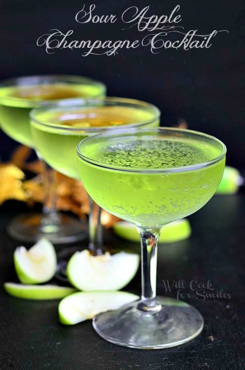 green Sour Apple Champagne Cocktail in a champagne glass on table with sliced green apples around it 