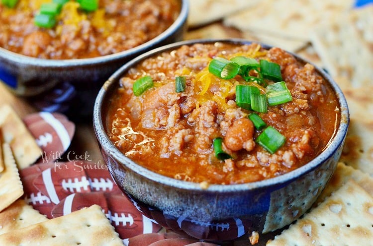 close up view of 2 small bowls of boozy chili on a blue platter with cracker scattered around bowls. Chili topped with shredded cheese and chives