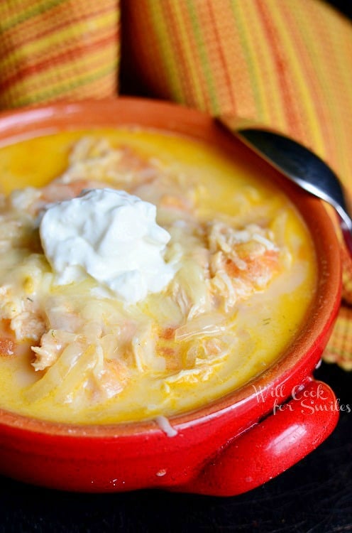 Creamy Chicken Enchilada Soup cu o lingură de smântână deasupra într-un bol roșu cu o lingură în stânga 