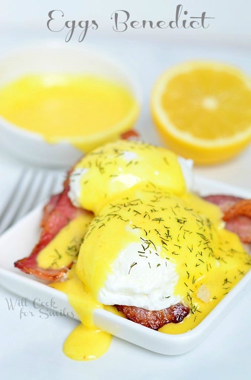 Hollandaise Sauce and Eggs Benedict over bacon on a white plate with a lemon to the top right and hollandaise sauce to the top left 