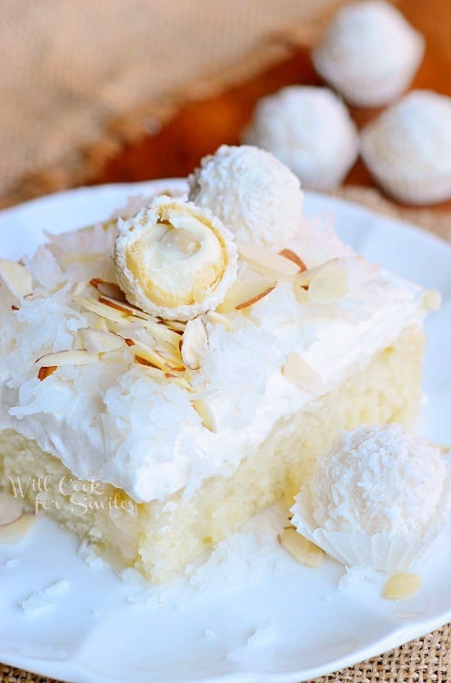 1 slice of rafaello poke cake coconut cake with white chocolate coconut cream and whipped topping on a small white plate on a brown burlap placemat on a wood table with Raffaello candy in background