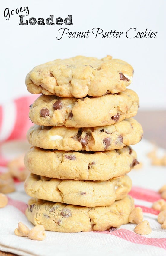 6 gooey loaded peanut butter cookies stacked on a white and red placemat with peanut butter chips scattered at base of stack