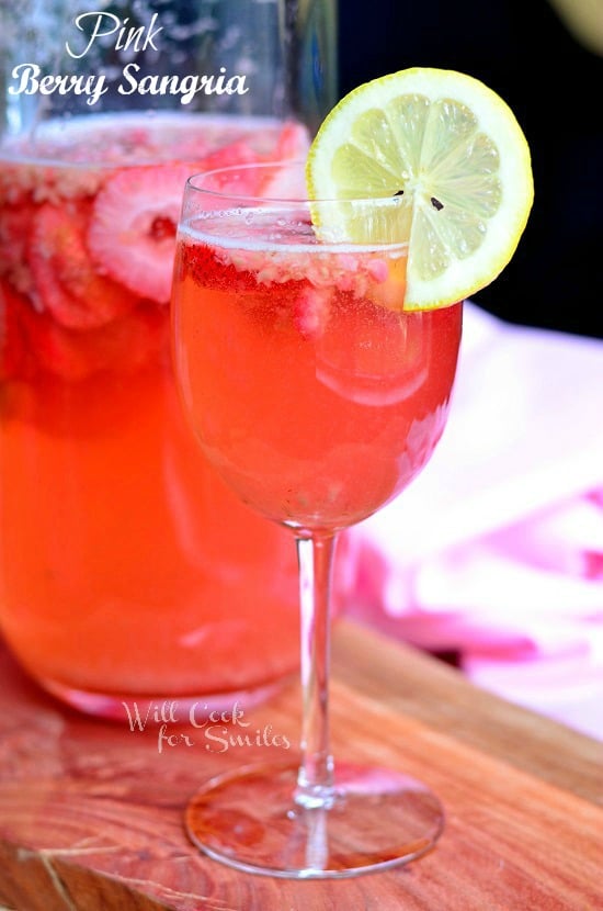 beverage jug filled with pink berry sangria on a wood plank with a wine glass filled with sangria in foreground