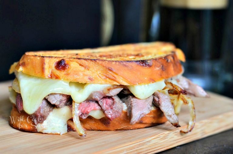 close up of steak and onion grilled cheese sandwich on a wood plank in front of a clear glass beer mug filled with stout beer and a bottle of beer in background