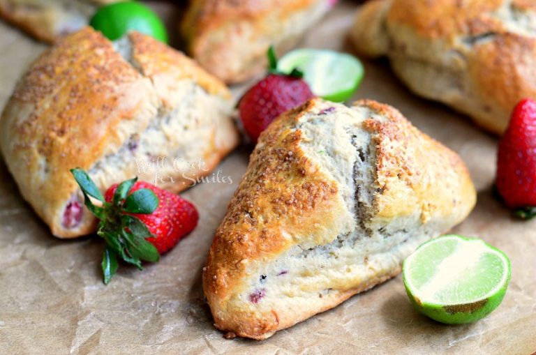 close up view of 1 strawberry key lime scones with strawberries and cut limes in foreground on a piece of brown wax paper and several more scones and fruit in background