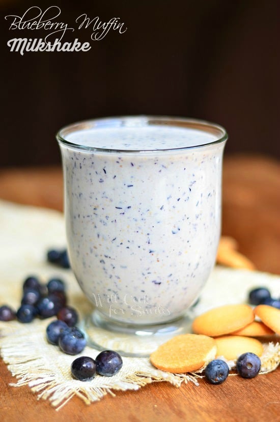 glass mug filled with blueberry muffin milkshake on a burlap cloth on wood table with blueberries and cookies scattered along the bottom of the glass