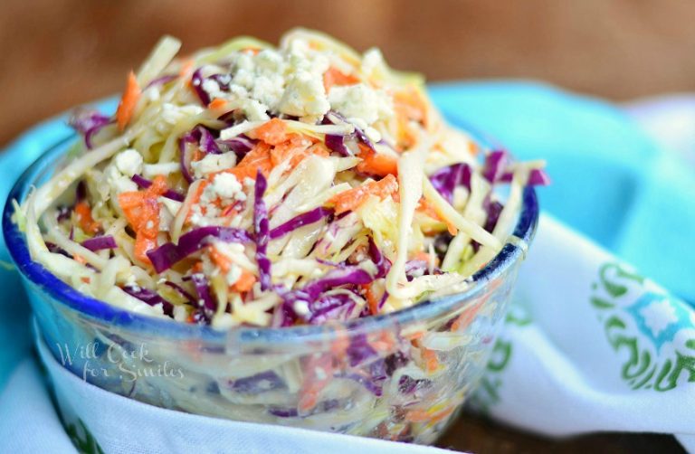 close up view of clear glass bowl filled with blue cheese coleslaw on a wood table with a white and green cloth in foreground and a blue cloth in background