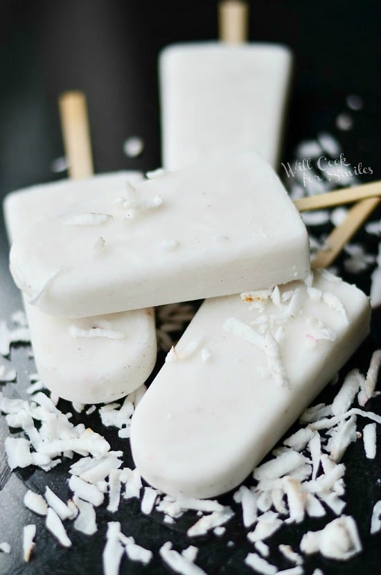 Coconut Vanilla Creamy Popsicles stacked up on a black table with coconut shavings around it 