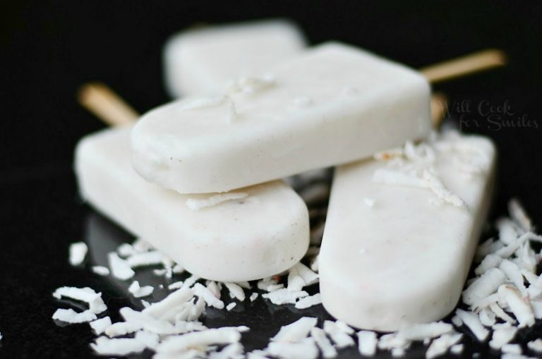 close up view of 3 coconut vanilla creamy pops on a black table with shredded coconut strewn across pops and table