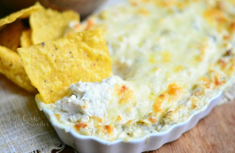 chip in dip in small white baking dish filled with chicken enchilada verde dip on a wood table with tan cloth to the left and a bowl of chips in background