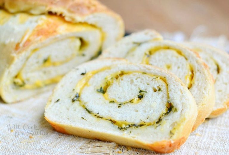 close up view of 2 slices of cheddar herb swirl bread on a brown placemat with the rest of the loaf behind in background