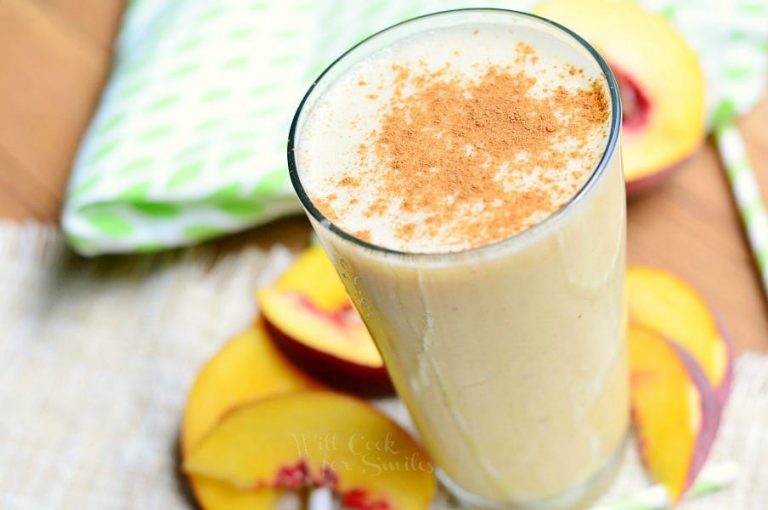 glass pint glass filled with peaches and cream breakfast smootie on a brown placemat on wood table with slices of peach at base of glass