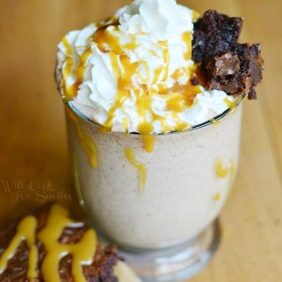 Glass mug filled with salted caramel brownie milkshake on a wooden table with a brownie in front of glass while both are drizzled with caramel