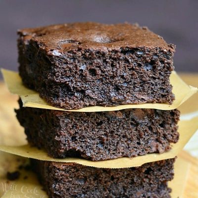 3 dark chocolate chocolate chip brownies stacked with wax paper in between each stacked on a wooden cutting board on a tand and white placemat on wood table