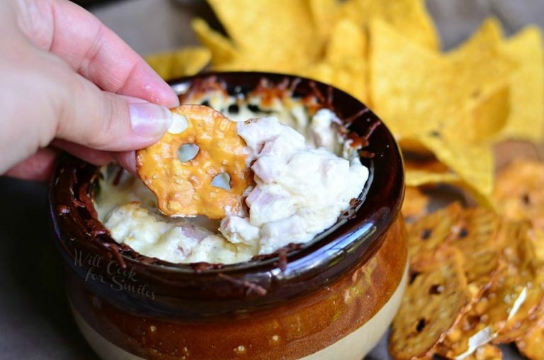 close up view of hand pulling out hot asiago ham dip portion on a pretzel chip out of a brown and tan crock with pretzels and chips scattered around base of crock while all sit on a blue cloth