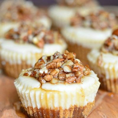 One cheesecake in foreground with 6 pecan mini cheesecakes lined up in 2 rows on a wood cutting board with maple syrup drizzled across the tops of the cupcakes