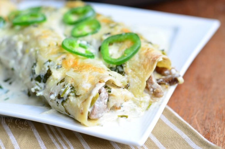 close up view of 2 steak enchiladas with jalapeno cilantro cream sauce on a white rectangle plate on a tan and white cloth on a wood table