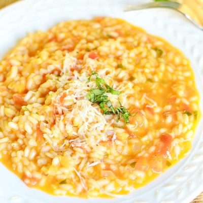 view from above of a white decorative rimmed bowl filled with creamy tomato basil risotto on a tan placemat on wooden table with leaves of basil as a garnish