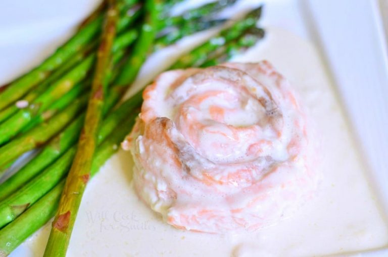 close up and viewed from above of white square plate with salmon filet molded into a rose design in white sauce with asparagus on a tan cloth on wood table with a sauce cup in background