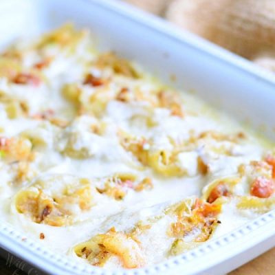 white baking dish filled with seafood alfredo stuffed shells on a tan placemat on wooden table with a silver serving spoon at bottom left of baking dish