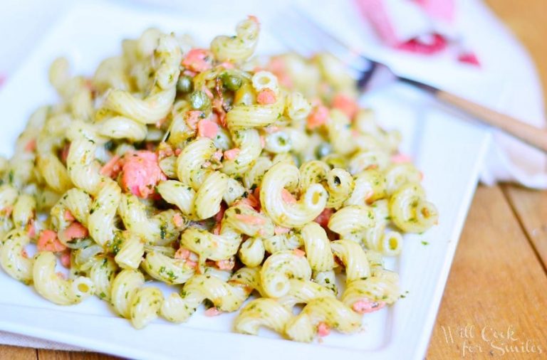 white square plate with smoked salmon pesto pasta and a fork with wooden handle on a wooden table with a white cloth with red stripes in the background