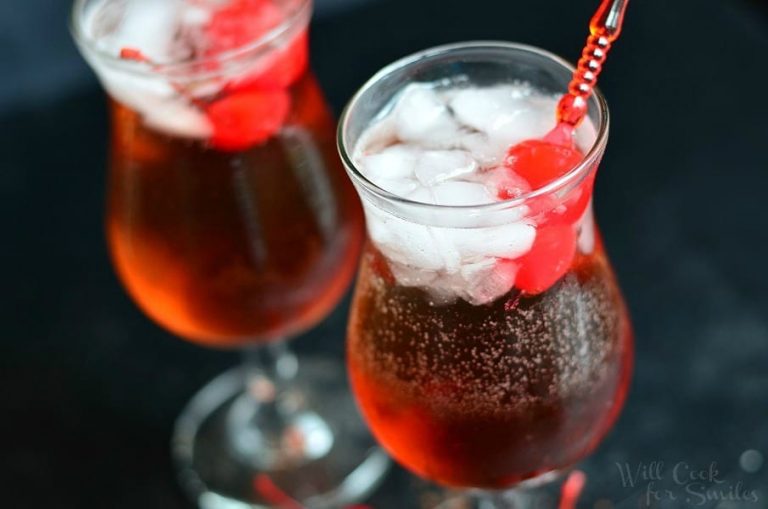 clsoe up view of 2 decorative wine glasses filled with "The pink lady raspberry cake coctail" on a black table with cherries at base of glass