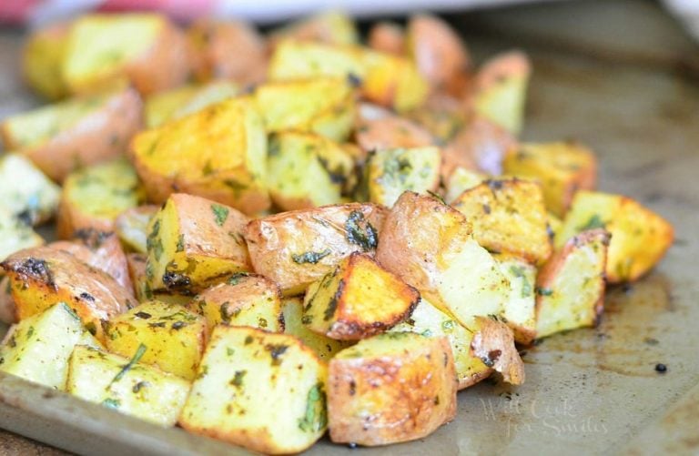 close up view of oven roasted potatoes cooked on a metal sheet tray presented on a wooden table with a white cloth with red stripes in background