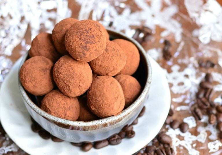 close up view of decorative white bowl filled above the brim with espresso chocolate truffles on a white plate with coffee beans strewn about the bottom while all sit on a wooden table