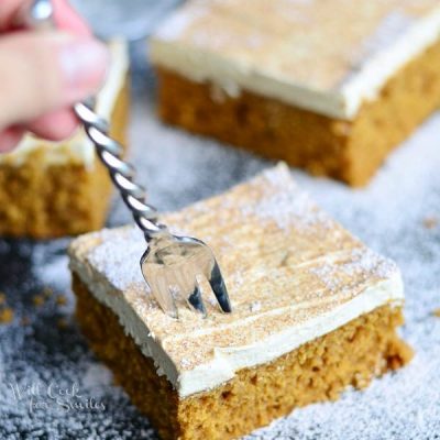 fork being held in cake square in foreground of 3 squares of gingerbread cake on a black table dusted with powdered sugar and a baking dish with more cake in the background