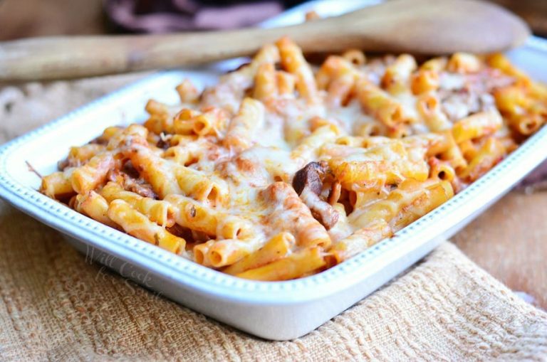 close up view of white baking dish with Itlian cheese steak baked ziti on a tan placemat with a wooden spoon spanning the corner of dish