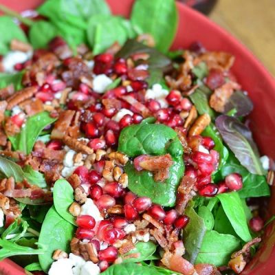 pomegranate goat cheese salad in a red baking dish on a tan placemat on wooden table with a bottle of dressing in background to the left