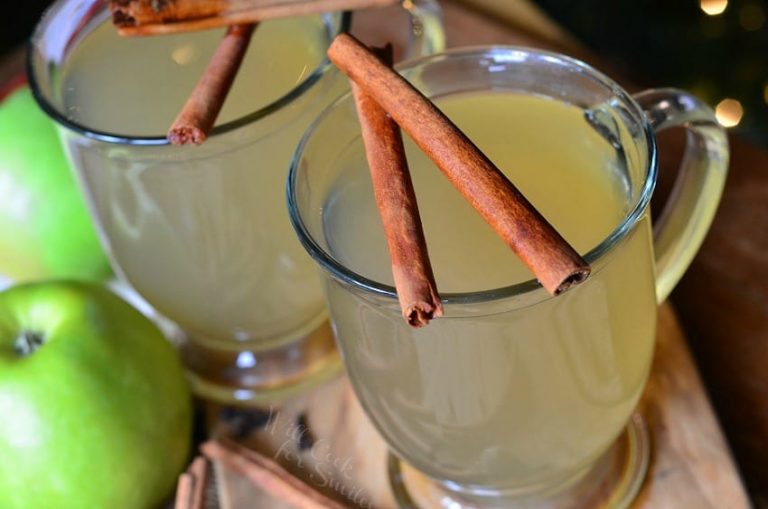 view from above of 2 glass mugs filled with crock pot mulled green apple cider and 2 cinnamon sticks laid across each glasses top rim as they both sit on a wooden table with more sticks at bottom of glasses.