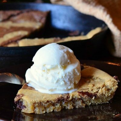 1 slice of vanilla bean marble skillet cookie on a black plate topped with a scoop of ice cream on a wooden table. The skillet with the rest of the cookie remains in the background with a tan cloth draped over the side.