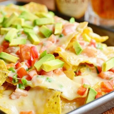 Close up view of Nachos with veggies and homemade nacho ale cheese on a silver baking pan on a wooden table with beer in the background